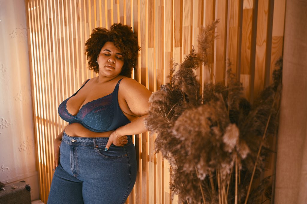 Beautiful stylish curvy oversize African black woman with afro hair posing in bra and jeans on wooden eco friendly background, body imperfection, body acceptance, body positive and diversity concept..